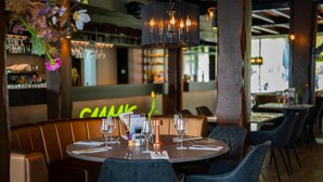 En restaurantindretning med runde borde, mørke stole og et barområde i baggrunden.
