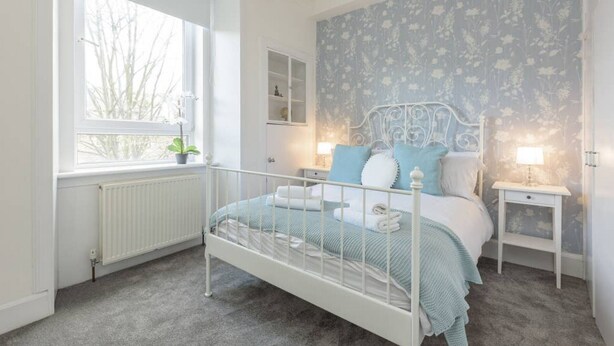 A bedroom with a white metal bed frame, a window with a view of bare trees, a radiator, and a floral wallpaper. - Balfour · Stylish and Comfortable City Centre Apartment (Edinburgh)