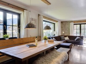 A spacious living area with a wooden bench, a sofa, and a dining table. There are large windows with curtains, a hanging light fixture, and a view of the outdoors. - Leogang Chalet Near Ski Lift & Bikepark (Leogang)