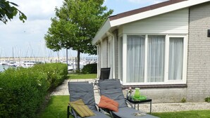 Une terrasse avec deux fauteuils et une petite table, une fenêtre avec des rideaux et, au loin, une vue sur des bateaux.