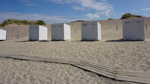 Une rangée de cabanes de plage blanches, dotées de portes, est installée sur une plage de sable, et une promenade en bois mène jusqu’à elles.