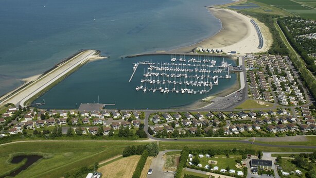 Vue aérienne d’une marina côtière où sont amarrés des bateaux, entourée de logements résidentiels et d’une plage de sable.