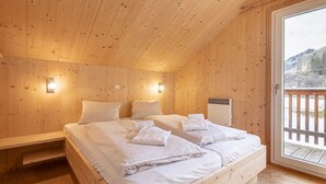 Een slaapkamer in een houten hut met een bed, twee nachtkastjes en een raam met uitzicht op een houten terras en heuvels.