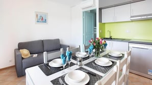 A dining area with a table set for four, a gray sofa, and a kitchen with white cabinets and a green backsplash. - Apartment in Grimaud With Pools (Grimaud)