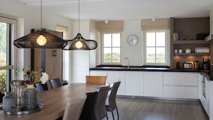 A modern kitchen with a wooden dining table, two pendant lights, a clock, and a view of the outdoors. - Friesland Villa With Fireplace (Delfstrahuizen)