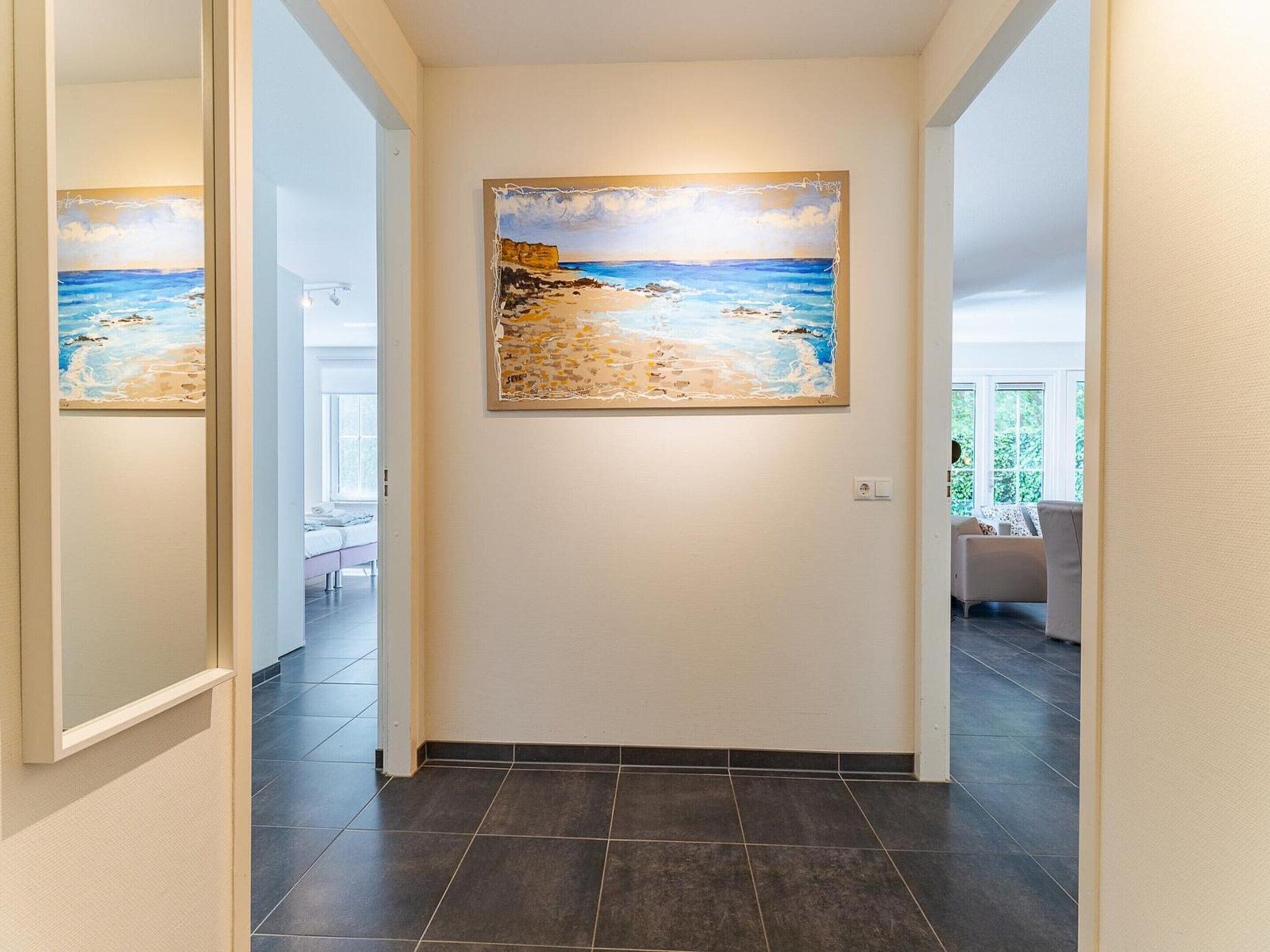 A hallway with a painting of a beach scene on the wall, a door, and a view into a room with a bed and a window.