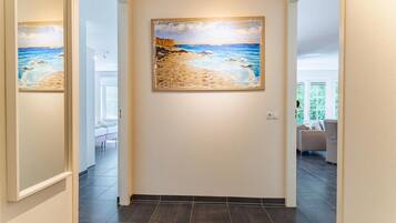 A hallway with a painting of a beach scene on the wall, a door, and a view into a room with a bed and a window.