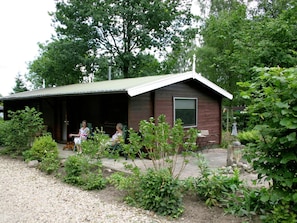 Een rustieke hut met een overdekte veranda, omgeven door weelderige begroeiing en een grindpad.