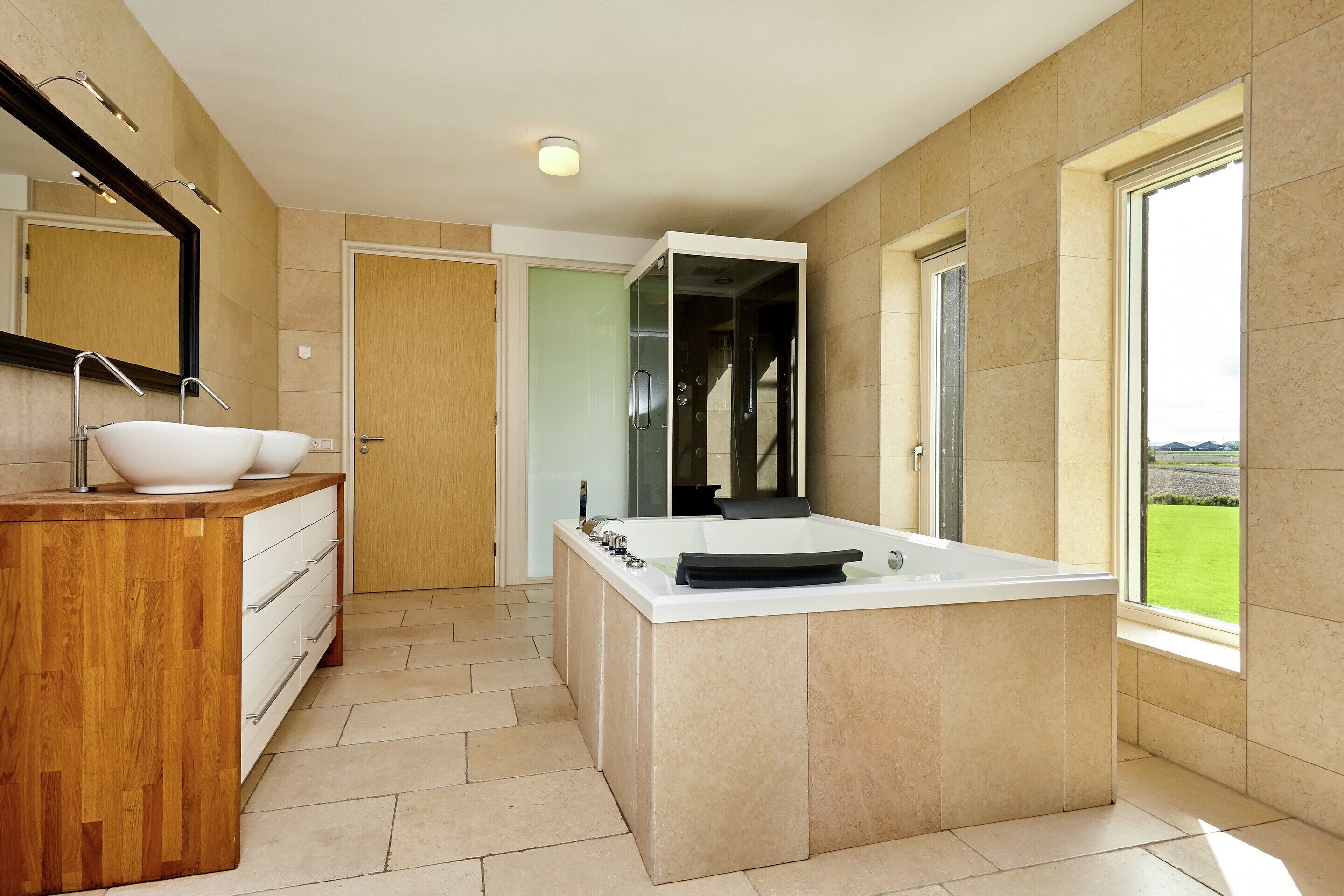 A modern bathroom with a large bathtub, a wooden vanity, a glass shower enclosure, and a view of the outdoors.