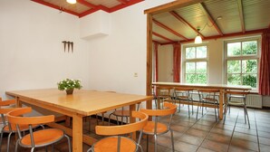 Zona pranzo con tavolo di legno e sedie arancioni, una pianta in vaso e vista sul verde attraverso ampie finestre.