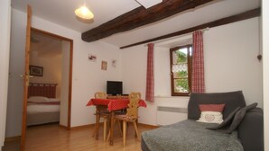 A room with a wooden floor, a grey sofa, a dining table with a red and white checkered tablecloth, wooden chairs, a television, and a window with red curtains. - Apartment in Riquewihr With Terrace (Riquewihr)