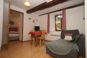 A room with a wooden floor, a grey sofa, a dining table with a red and white checkered tablecloth, wooden chairs, a television, and a window with red curtains. - Apartment in Riquewihr With Terrace (Riquewihr)