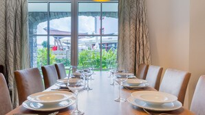 Mesa de comedor dispuesta para seis personas con sillas, lámpara colgante y vista a un área exterior con sombrillas y un cartel de Coca-Cola.