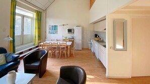 A spacious living area with a dining table, chairs, a sofa, a kitchen with appliances, and a large window with curtains.