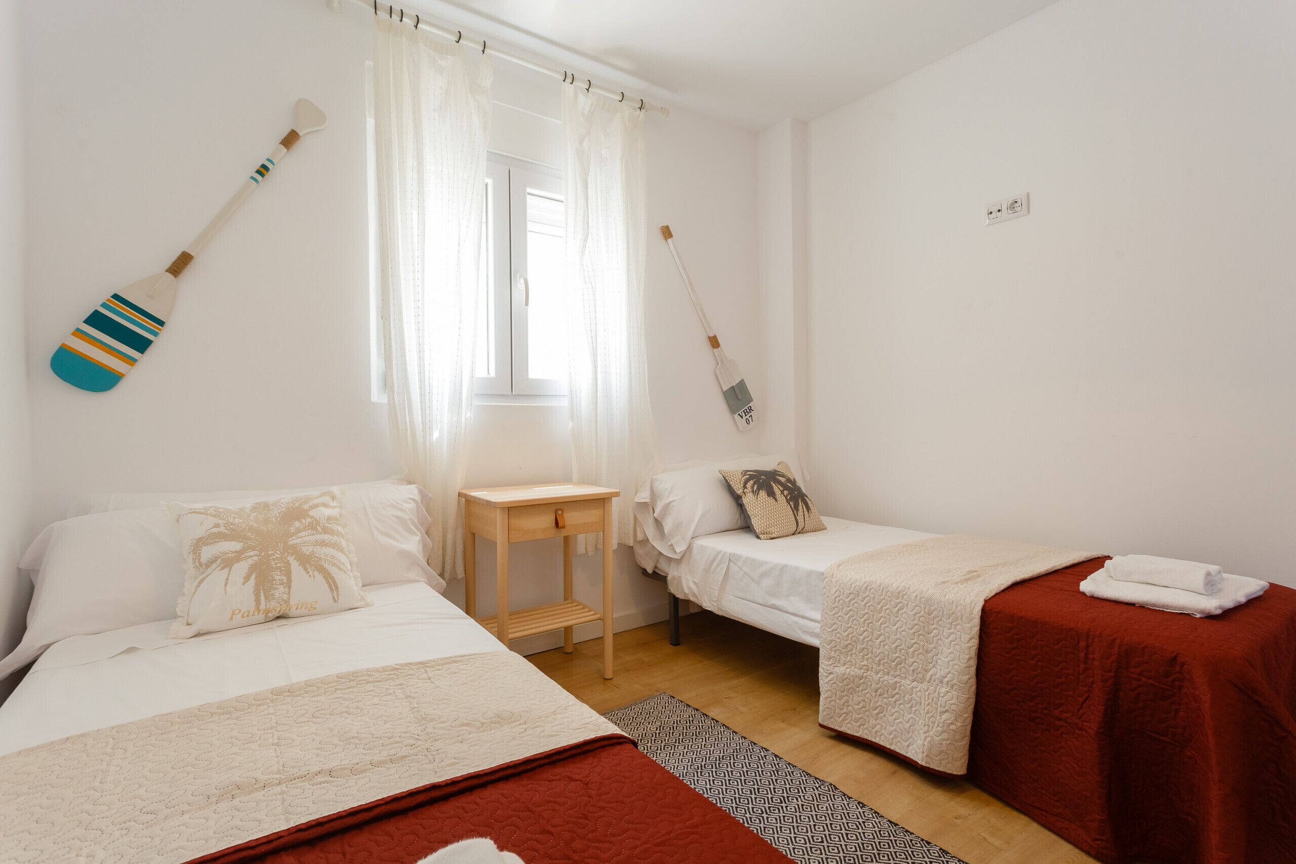 A hotel room with two beds, a small table, a window with sheer curtains, and a wall-mounted light switch.