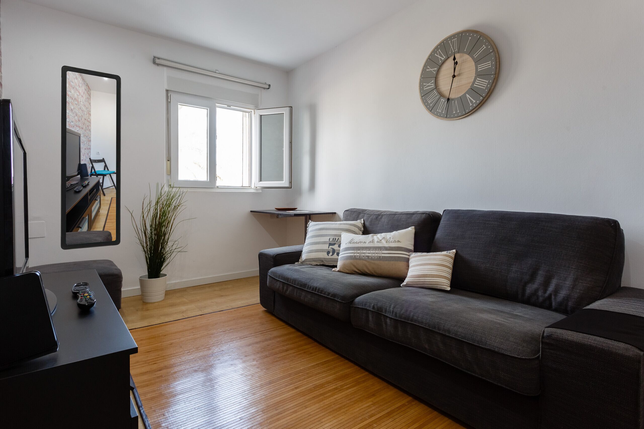 A living room with a dark grey sofa, a wooden floor, a large wall clock, a mirror, and a potted plant.