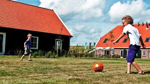 Zwei Kinder spielen mit einem Fußball auf einer Wiese, im Hintergrund ein Gebäude mit rotem Dach.