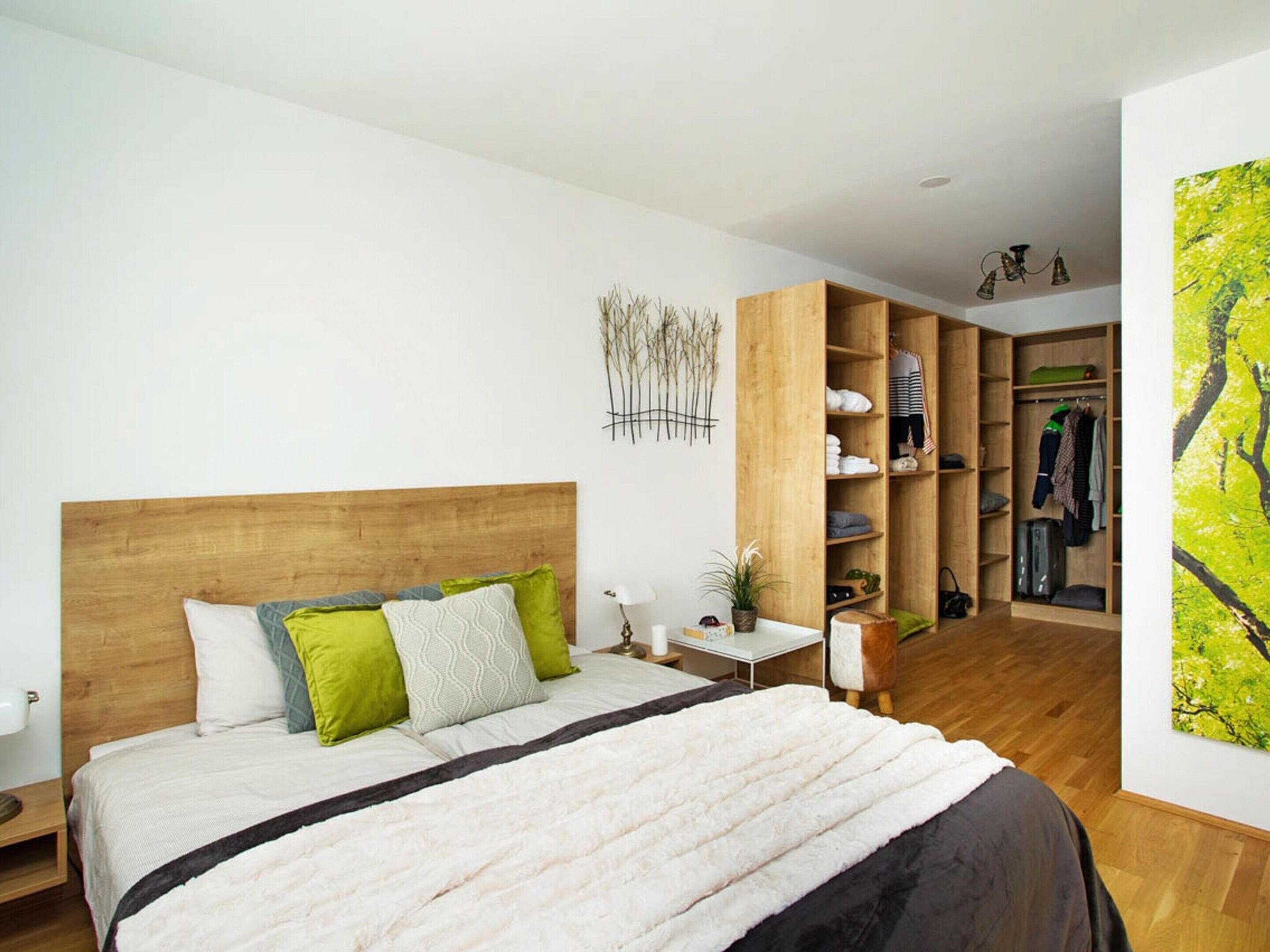 A bedroom with a wooden headboard, a bed with a white and black duvet, green and white pillows, a bedside table with a lamp, a small plant, and a wooden wardrobe with shelves.