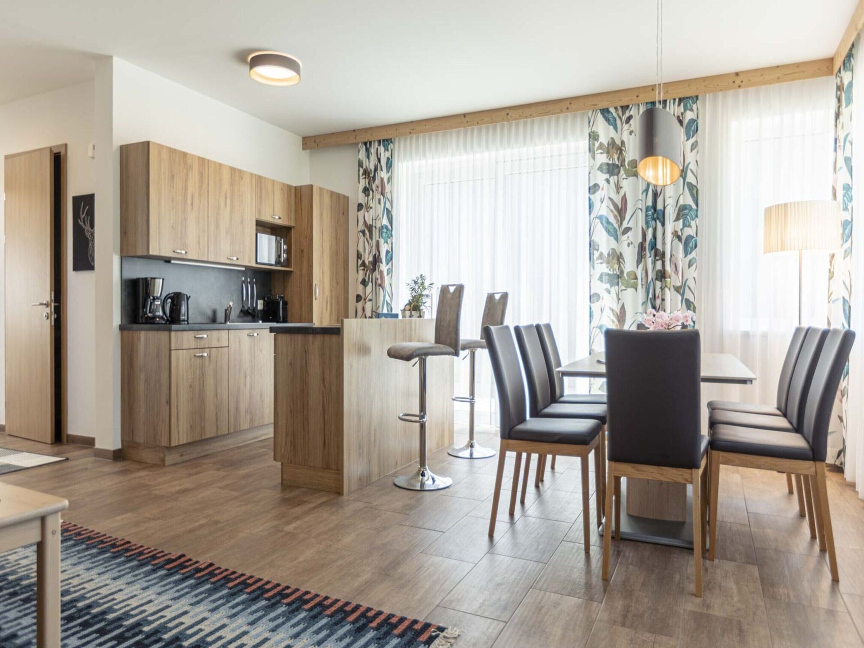 A modern kitchen with wooden cabinets, a dining area with a table and chairs, and a large window with curtains.