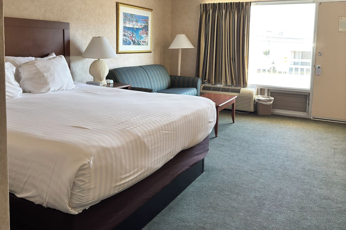 Ein Hotelzimmer mit einem großen Bett, einer Couch, einem Schreibtisch, einer Lampe, einem Gemälde und einem Fenster mit Vorhängen.