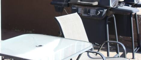 Un patio al aire libre con una mesa de vidrio, sillas de metal, una parrilla y una barandilla de metal.