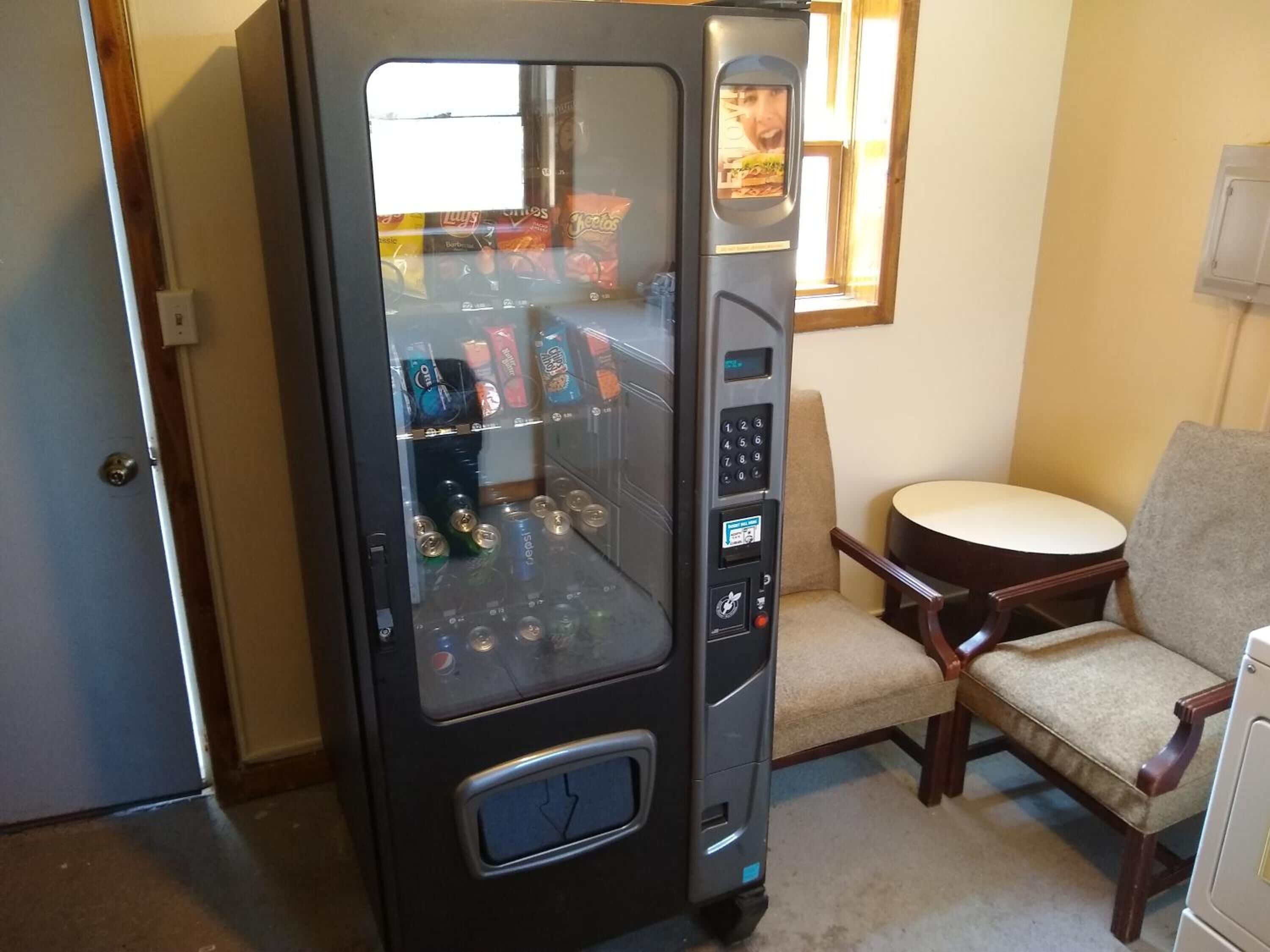 Máquina expendedora con diferentes aperitivos y bebidas, ubicada en una sala con una silla y una pequeña mesa.