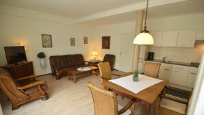 Una sala de estar con un sofá, sillas, una mesa de centro y un televisor. Hay una mesa de comedor con sillas y una zona de cocina con armarios y un lavabo.