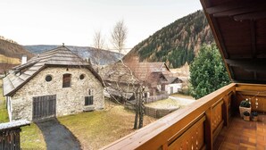 A stone building with a sloped roof, a wooden balcony, and a view of a village with houses and a forested mountain in the background. - Holiday Home in Rennweg Near Ski Slopes (Rennweg am Katschberg)