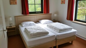 Un lit double avec du linge de lit blanc et une tête de lit en bois, deux tables de chevet avec des lampes et une fenêtre avec des rideaux rouges.