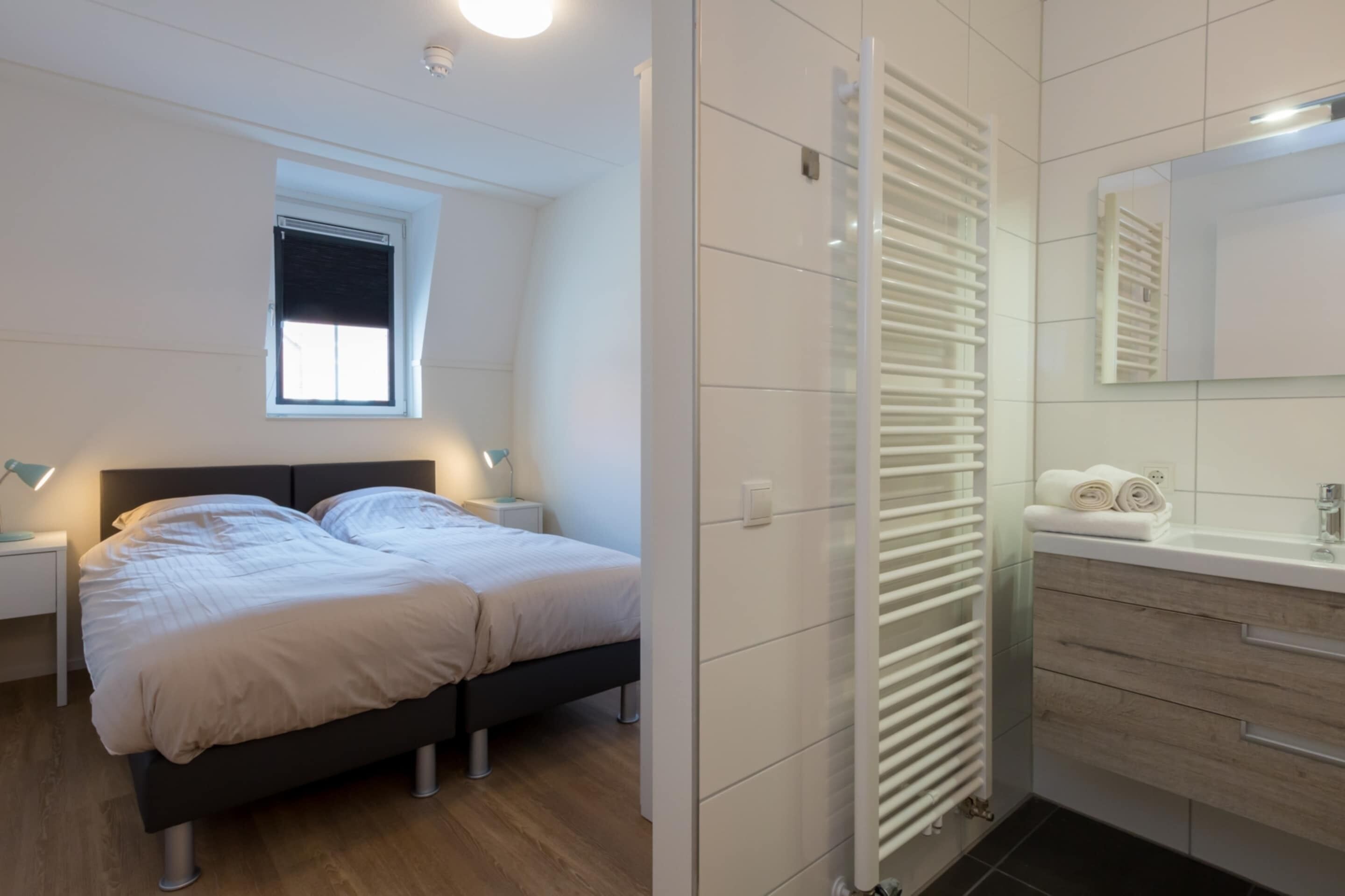 A modern bedroom with a bed, bedside tables, a window, a radiator, and a bathroom with a mirror and towel rack.