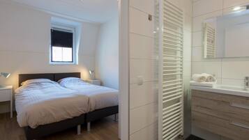 A modern bedroom with a bed, bedside tables, a window, a radiator, and a bathroom with a mirror and towel rack.