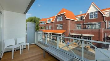 A balcony with white chairs, a wooden floor, and a view of red brick buildings with orange roofs.
