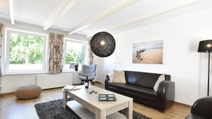 A modern living room with a black leather sofa, a wooden coffee table, a round ottoman, and a large black pendant light. - Cosy Holiday Home With Large Garden (Zonnemaire)