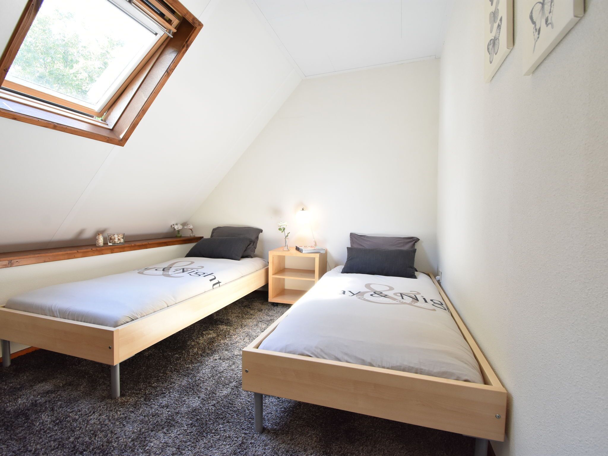 A small, well-lit room with two wooden beds, a skylight, and a small bedside table with a lamp.