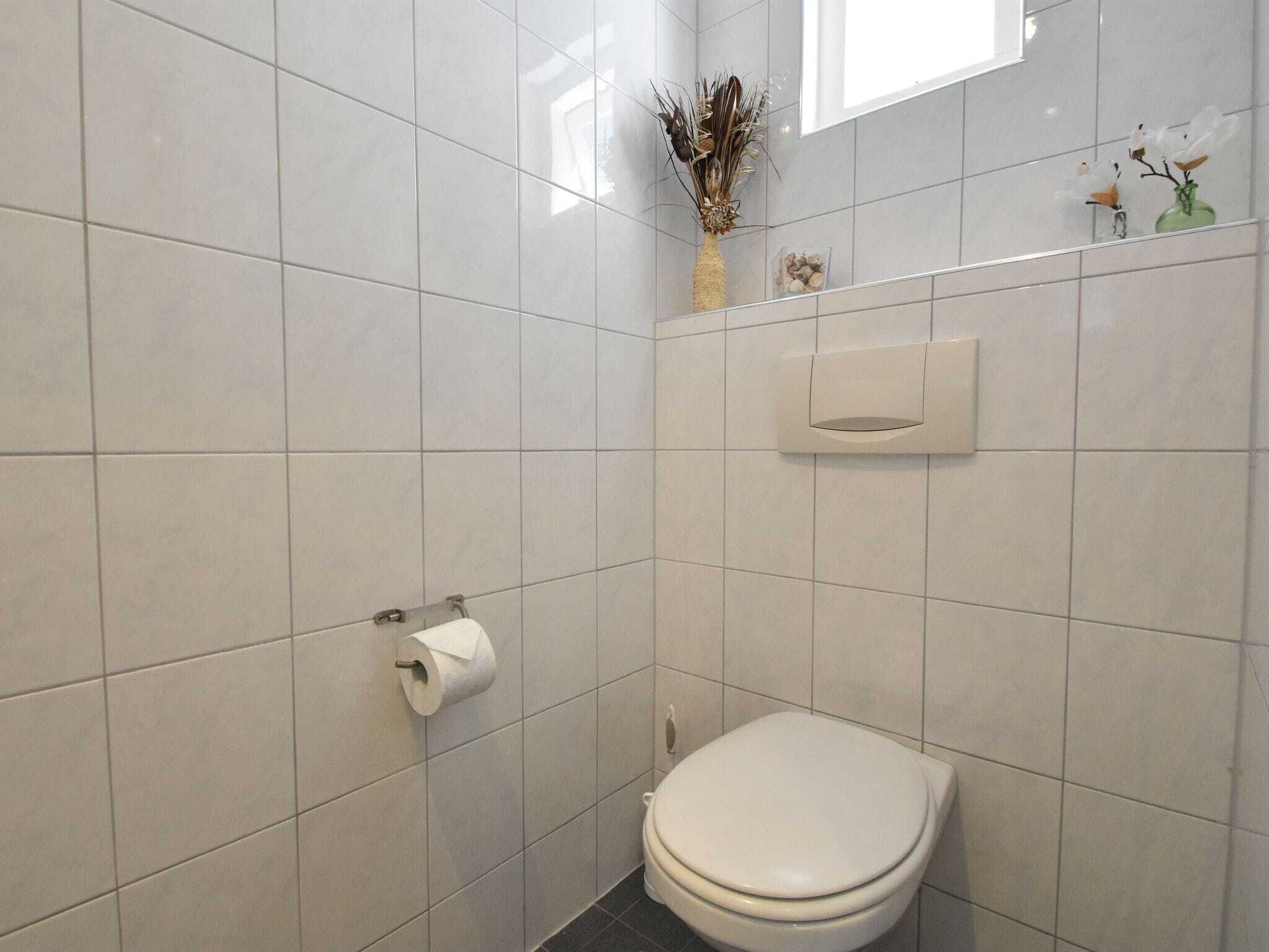 A small, modern bathroom with white tiles, a toilet, a paper towel dispenser, and a small vase with dried flowers on the wall.