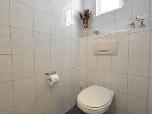A small, modern bathroom with white tiles, a toilet, a paper towel dispenser, and a small vase with dried flowers on the wall.