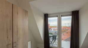 Huone, jossa on kalteva katto, ikkuna verhoilla, patteri ja näkymä ulkona oleviin rakennuksiin.