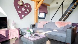 A modern living room with a grey sofa, a pink ottoman, and a TV on a pink dresser. A heart-shaped wall decoration and a staircase are visible. - Holiday Home in Alphen-chaam With Terrace (Alphen)