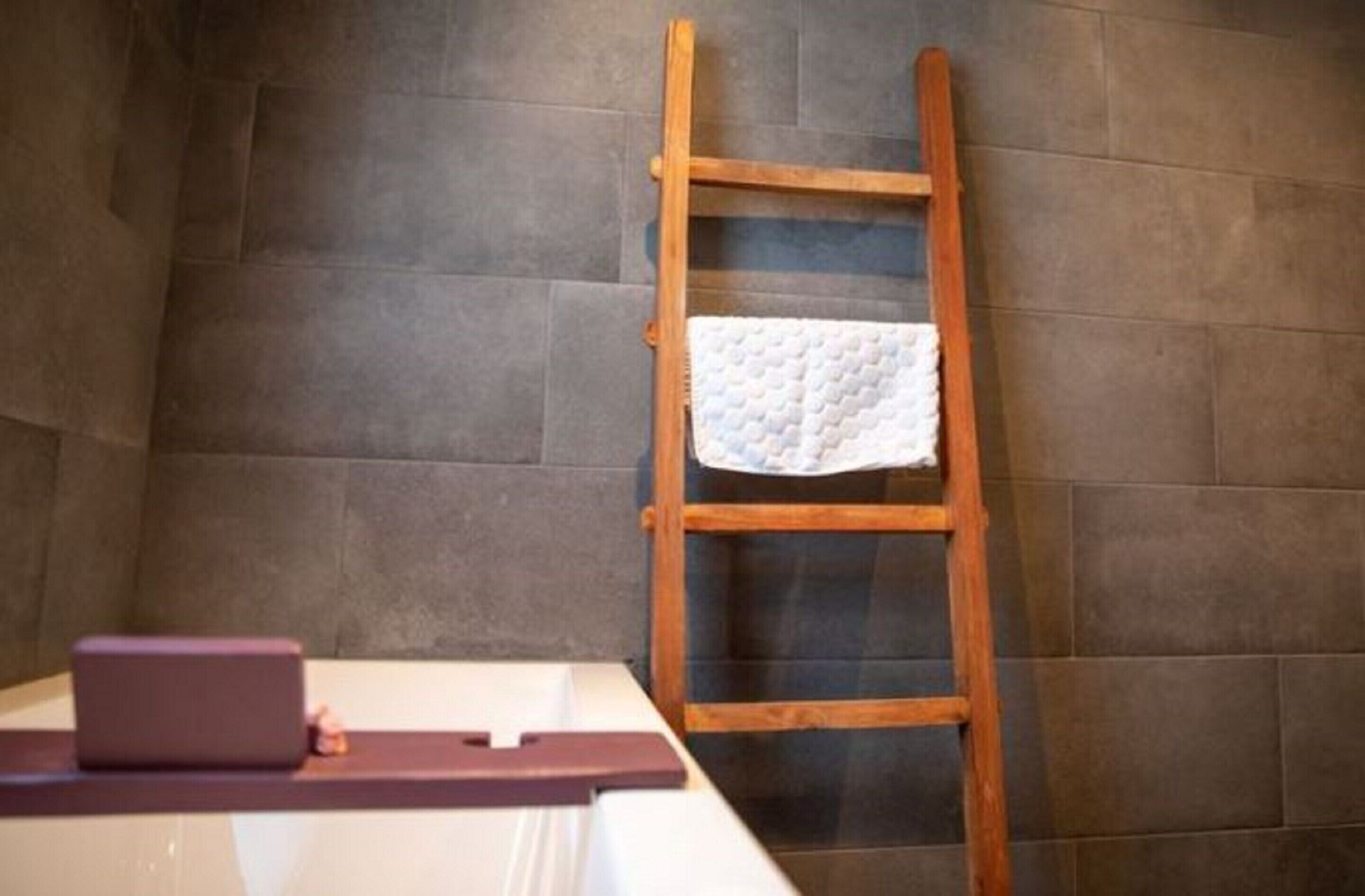 Um banheiro com uma escada de madeira, uma toalha branca pendurada nela e um objeto roxo no chão.
