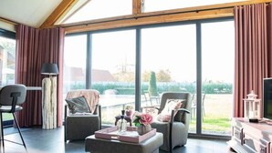 A spacious living room with large windows, a sofa, a coffee table, and a television. - Holiday Home in Alphen-chaam With Terrace (Alphen)