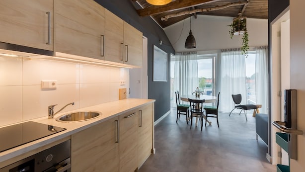 A modern kitchen with a built-in oven, a sink, and wooden cabinets. There is a dining area with a table and chairs, and a large window with curtains. - Holiday Home in Koudekerke Near Seabeach (Koudekerke)
