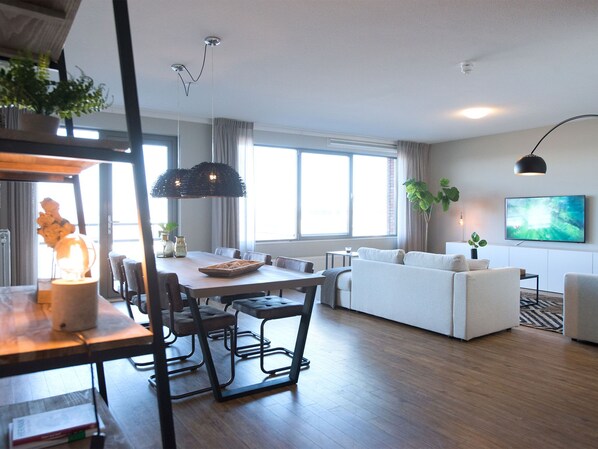 A modern living room with a sofa, dining table, and chairs. There is a large window providing natural light and a view of the outdoors. - Sea-view Apartment With Balcony (The Hague)
