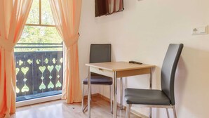 Habitación con una mesa, dos sillas, una ventana con cortinas naranjas, un cuadro en la pared y vista a zonas verdes.