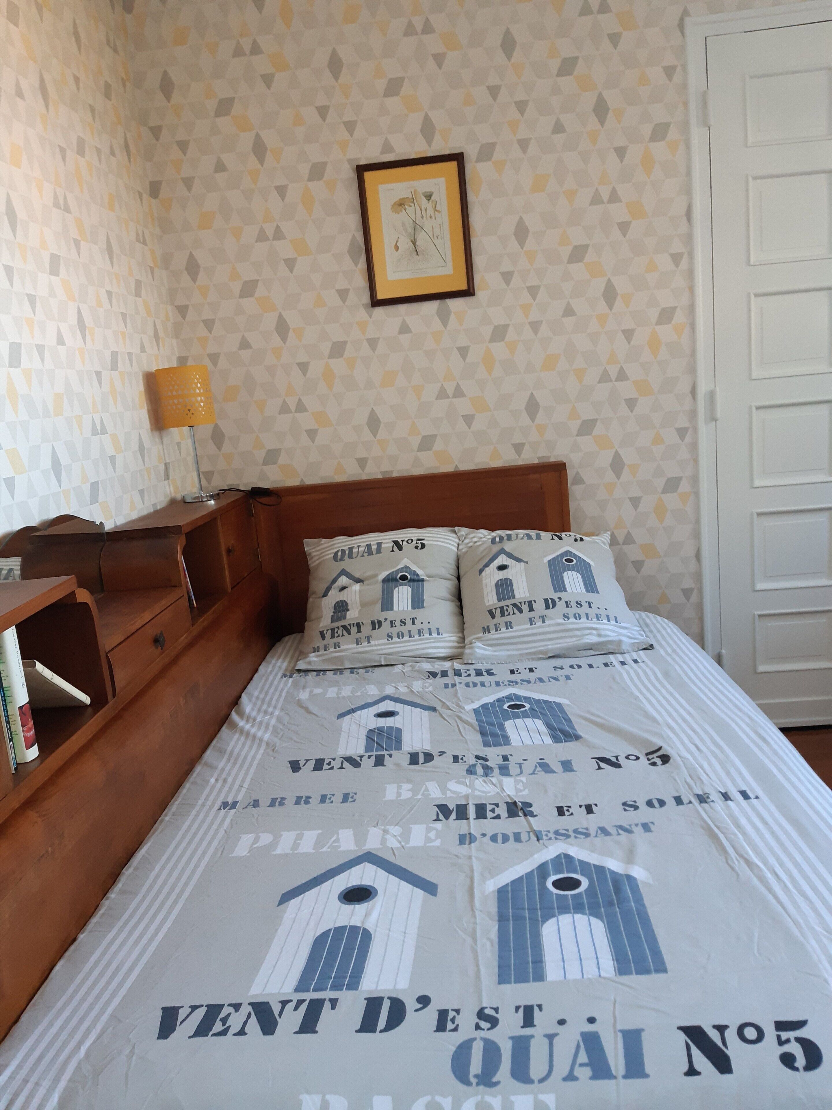 A bedroom with a bed, pillows, a nightstand, a lamp, a framed picture on the wall, and a patterned wallpaper.