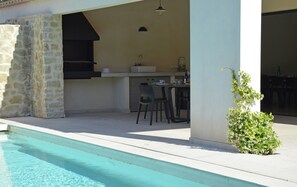 A poolside area with a stone wall, a barbecue grill, a table with chairs, and a small plant. - Villa in Malaucene With Private Pool (Malaucene)