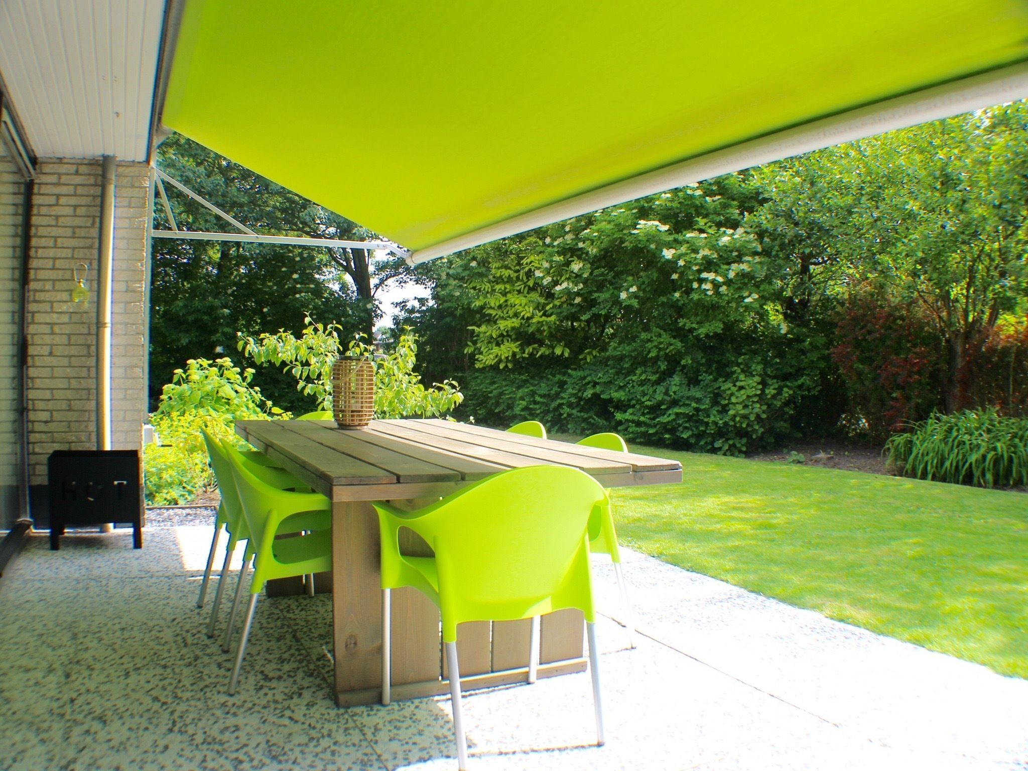 An outdoor patio with a wooden table and bright green chairs, surrounded by a covered area and a view of a lush garden.