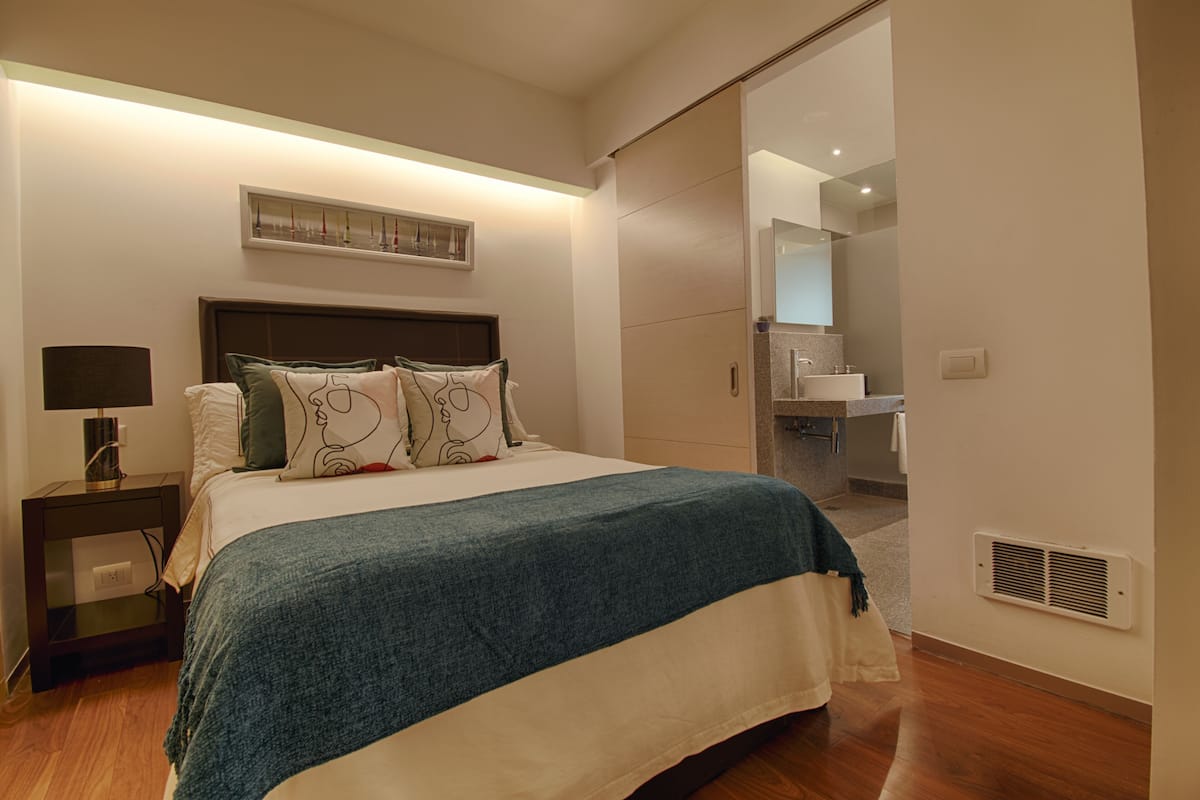Une chambre à coucher bien rangée, avec un lit, une table de chevet et une salle de bain visible à travers une porte ouverte.