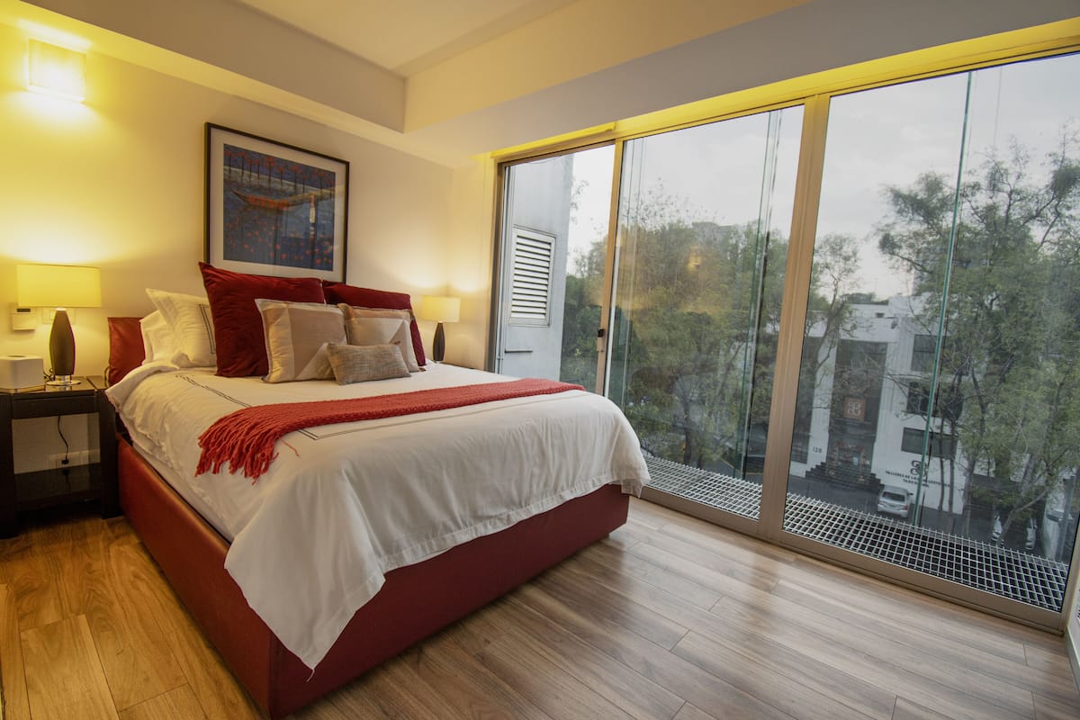 A modern bedroom with a large bed, a nightstand, a window with a view of buildings and trees, and a ceiling light.