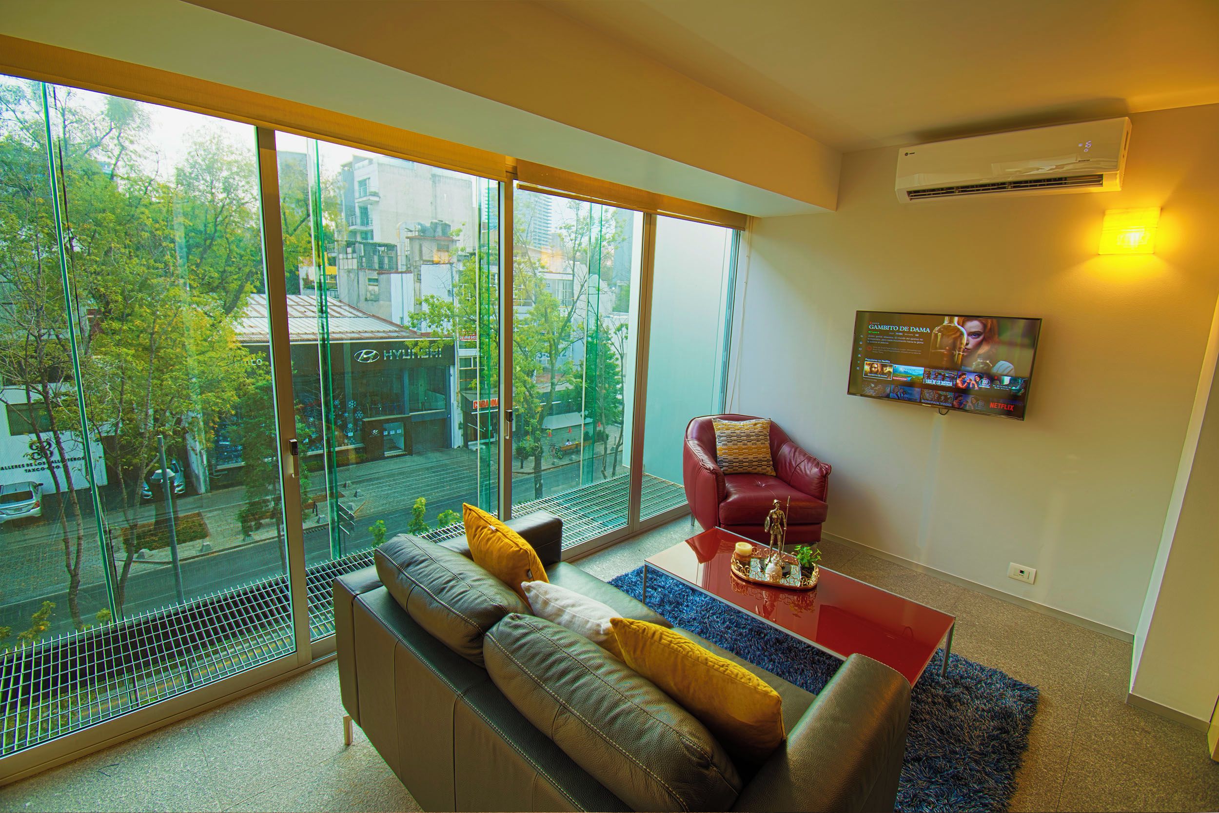 A modern living room with a large window, a red armchair, a sofa with cushions, a coffee table, and a mounted TV.
