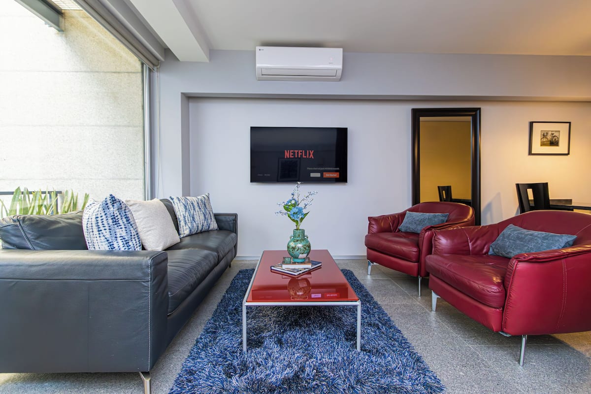 A modern living room with a grey sofa, a red leather sofa, a coffee table, and a TV mounted on the wall.